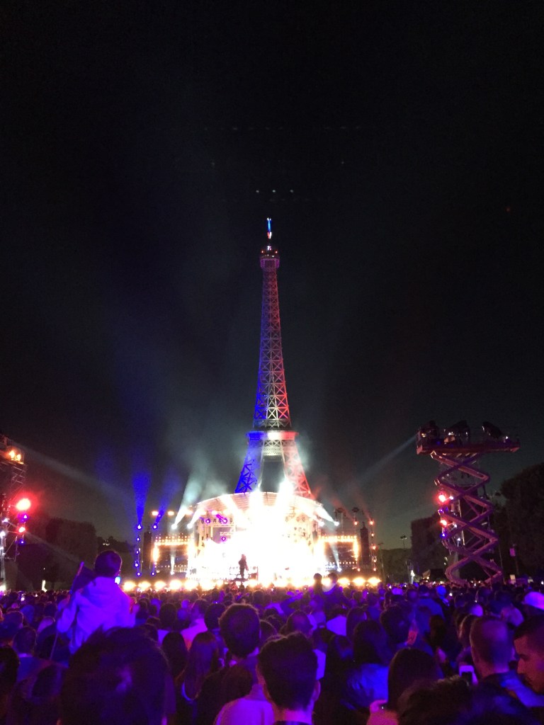 Eiffel Tower lit up at nighttime with the colors of the French flag (blue, white, red) and a crowd of people in front to celebrate Bastille Day.