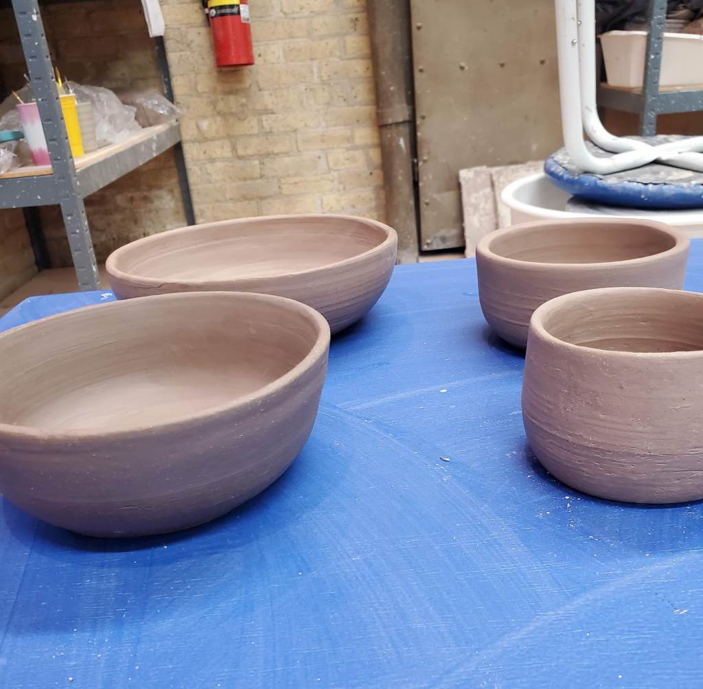 Two large clay bowls and two small clay bowls sit on a worktable in a pottery studio. They are ready to dry out and then be fired in a kiln.
