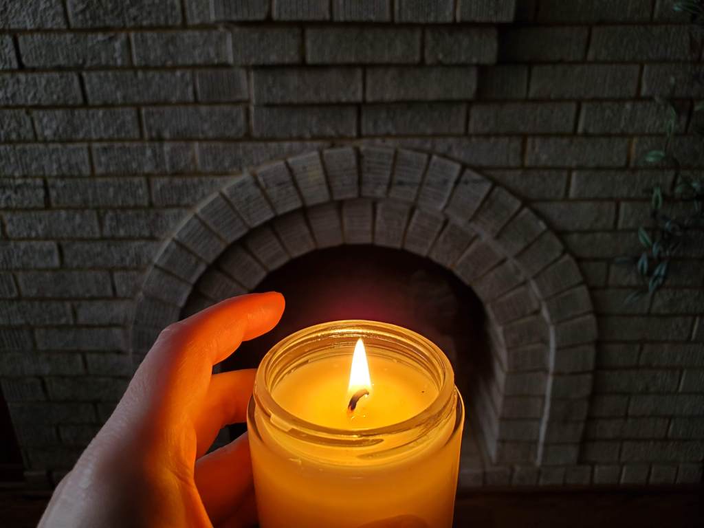 foreground shows a hand holding a lit candle with a brick fireplace in background