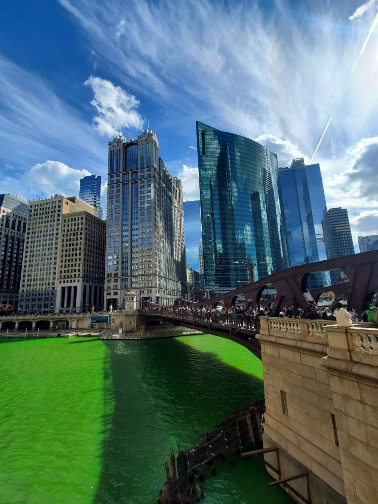 Bright blue sky and skyscrapers in the background as a crowd gathers along a bridge to look down at the neon green water of the Chicago River