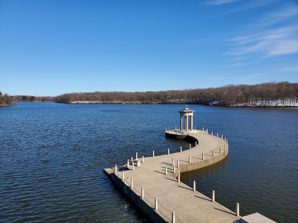Blue skies over a calm blue lake with a white concrete path pier cutting through the water and leading to a gazebo at end of pier